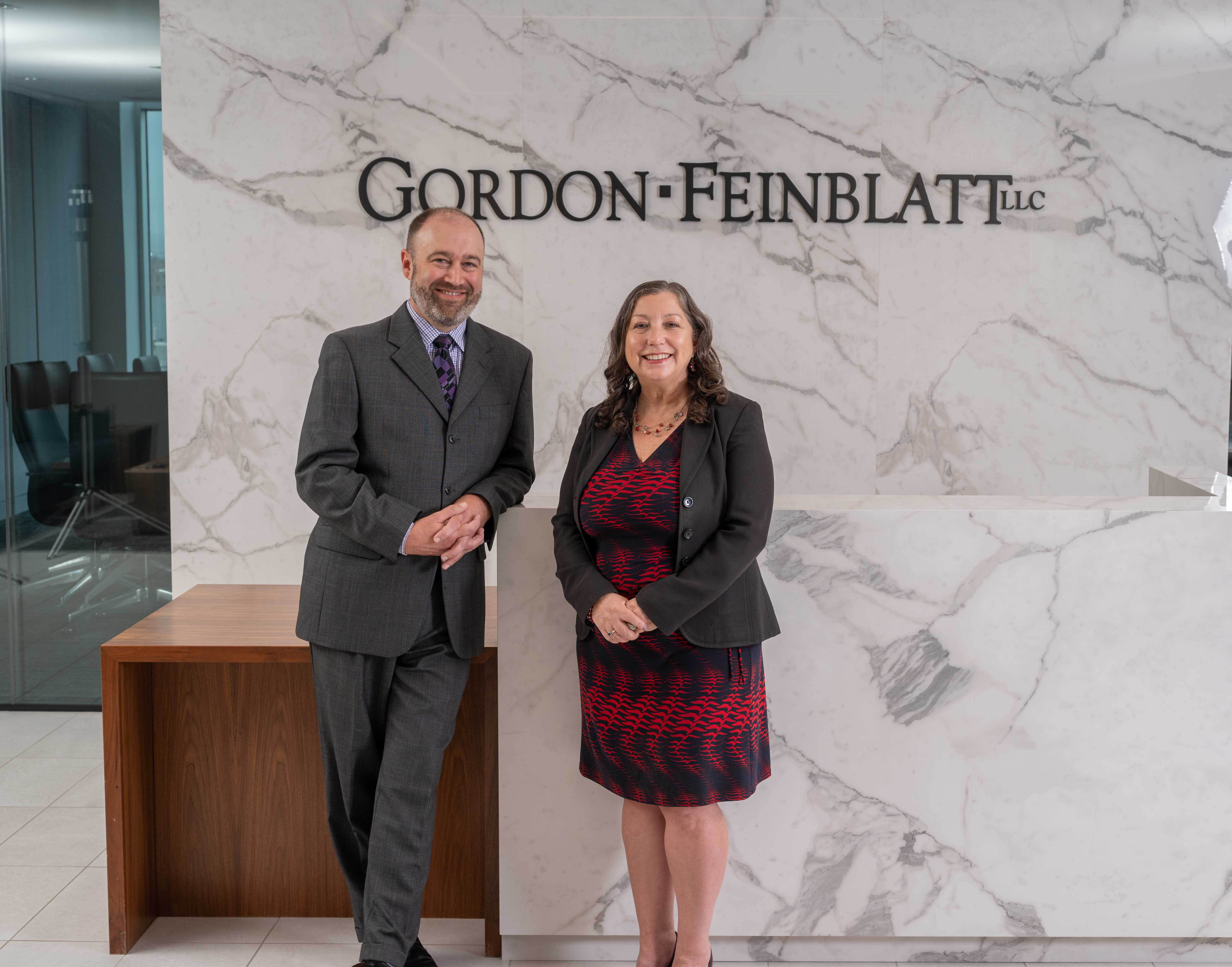 Photo of our CEO and Chair, Michele Walsh, standing in a red and black dress with a black blazer, alongside our Managing Member, Todd Chason, who is wearing a light gray suit with a purple shirt and a purple and black patterned tie.